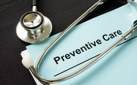 Macro close-up photo of a medical checklist document highlighting the printed words 'Preventive Care', with the chrome head of a stethoscope resting in sharp focus right beside the text.