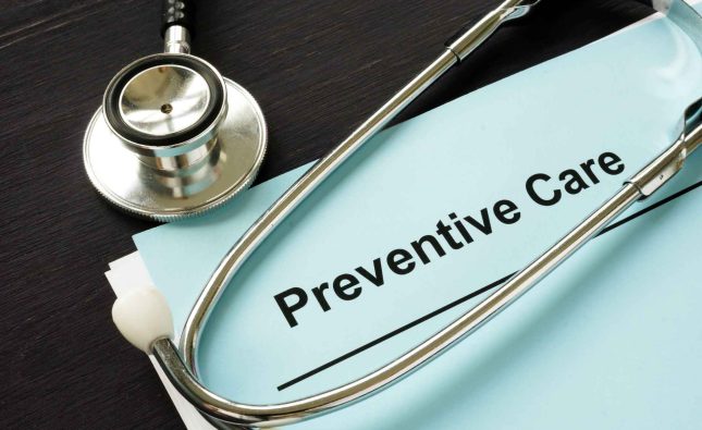 Macro close-up photo of a medical checklist document highlighting the printed words 'Preventive Care', with the chrome head of a stethoscope resting in sharp focus right beside the text.